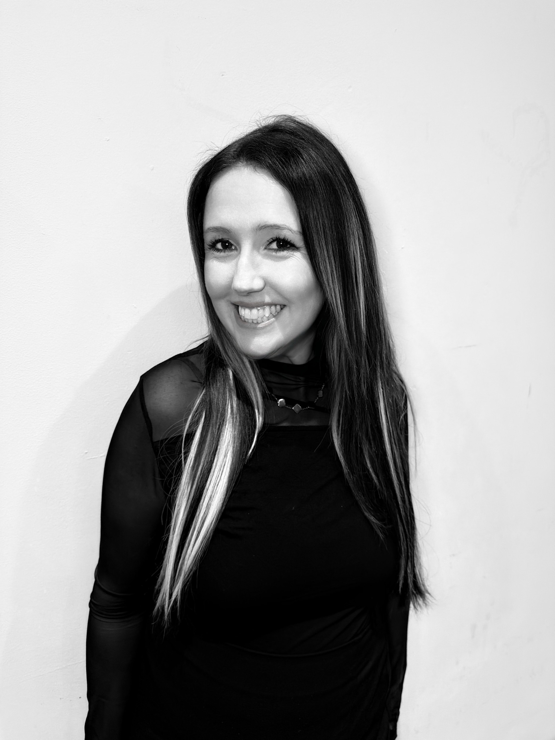 Portrait of a smiling woman with long dark hair wearing a black top against a pale wall