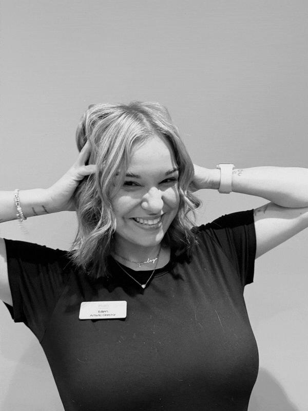 Smiling woman with short blonde hair wearing a name badge and black shirt hands resting on her head against a plain background