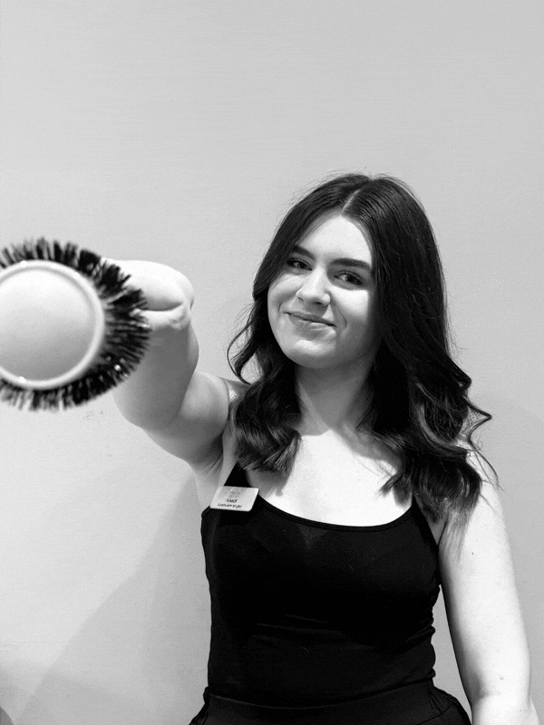 Smiling young woman in a black top at a salon holding a round hairbrush toward the camera
