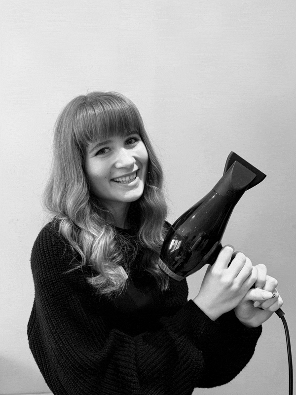 Smiling woman with wavy hair wearing a dark sweater holding a black hair dryer as if preparing to use it